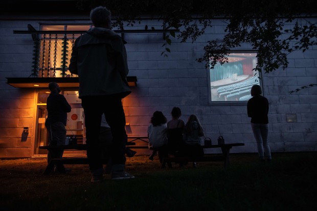 Rétroprojection vidéo présentée dans une fenêtre de l'édifice de La Filature. Quelques personnes sont assises sur un banc et d'autres sont debout.