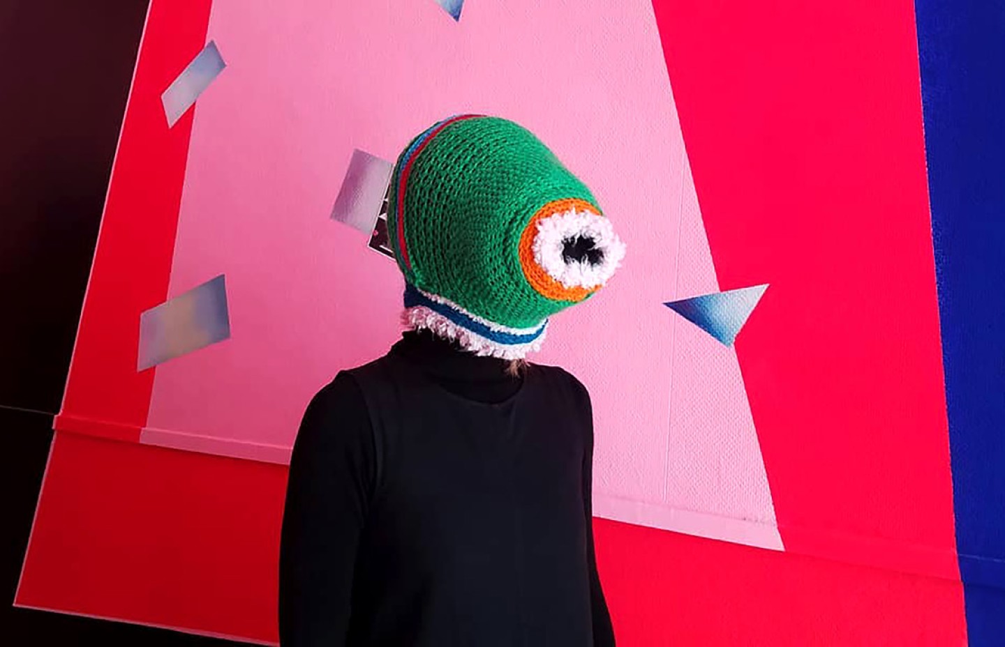 Une personne portant un masque crocheté vert coloré et des vêtements noirs est debout devant un fond coloré rose, rouge et bleu avec des motifs géométrique