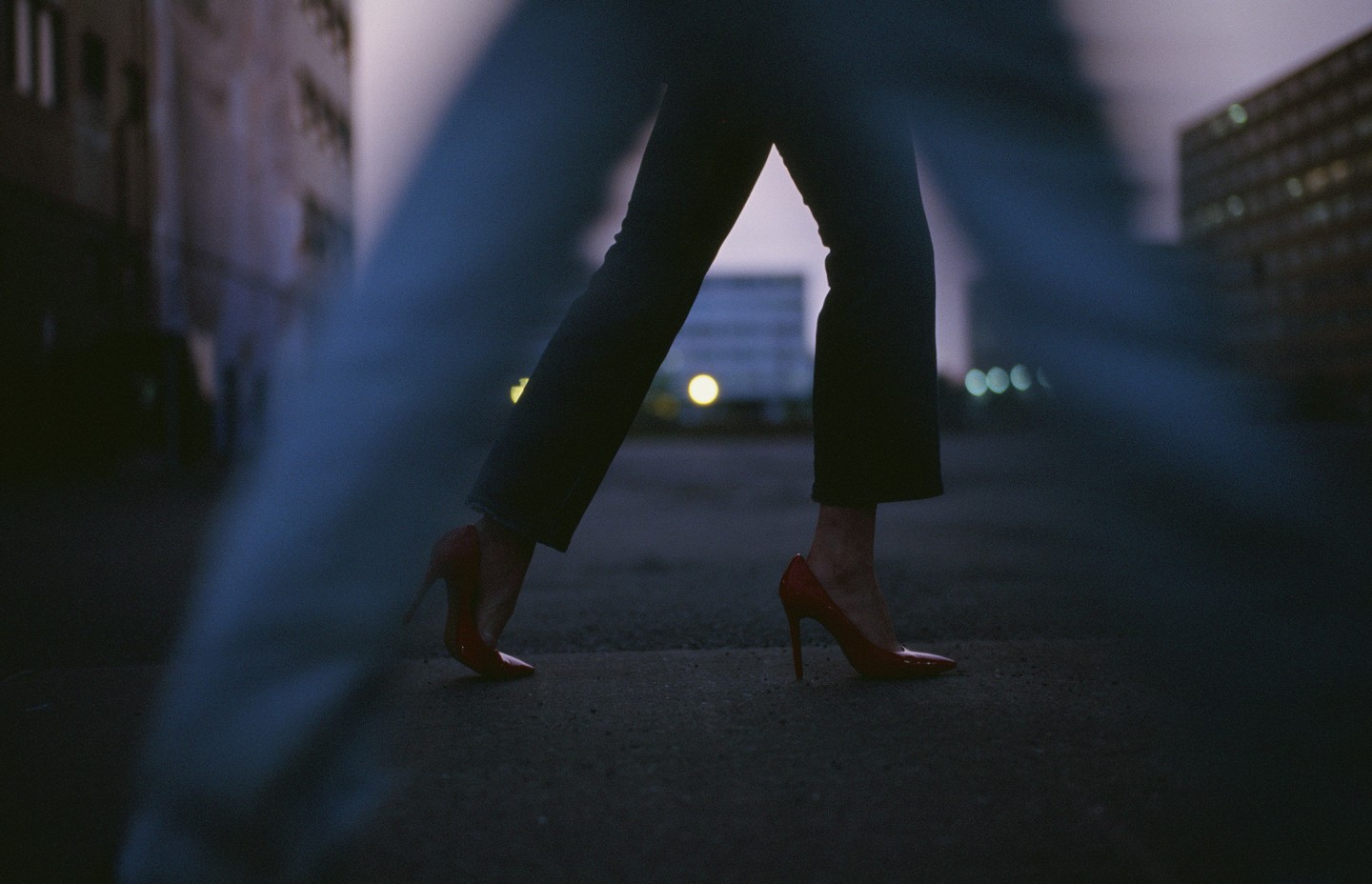 Une photographie d'un plan rapproché au niveau des jambes de deux personnes qui marchent dans la rue. Au premier plan, les jambes sont floues. Au deuxième plan, la personne qui marche porte des souliers à talon hauts rouge.