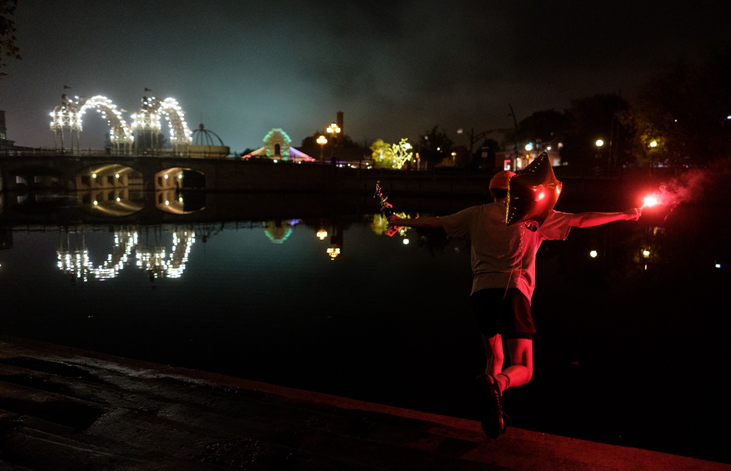 une personne sur le bord d'une rivière avec un ballon en forme d'étoile, une torche rouge à la main et un trophée. Il fait nuit et il y a un pont éclairé par des lumières