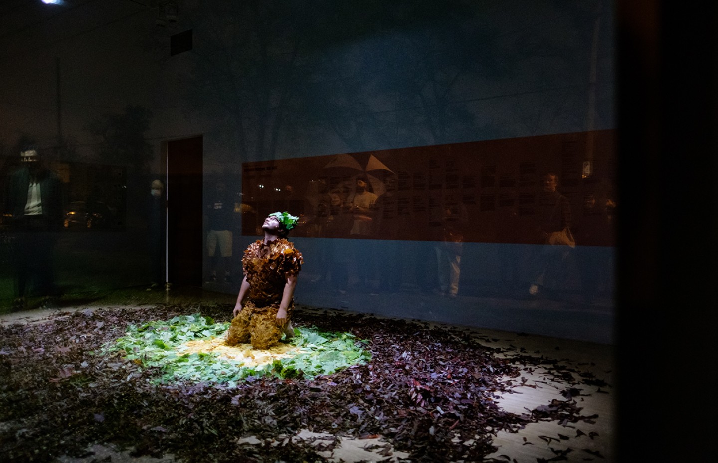 Dans cette image, on voit une performance artistique dans un espace sombre. La personne au centre, habillée en un costume fait de feuilles et de matières naturelles, est accroupie sur un tapis de feuilles et de végétaux, avec une lumière focalisée sur elle, illuminant la scène de manière dramatique. Autour d'elle, on distingue des spectateurs observant la scène à travers une vitre, reflétés dans le verre