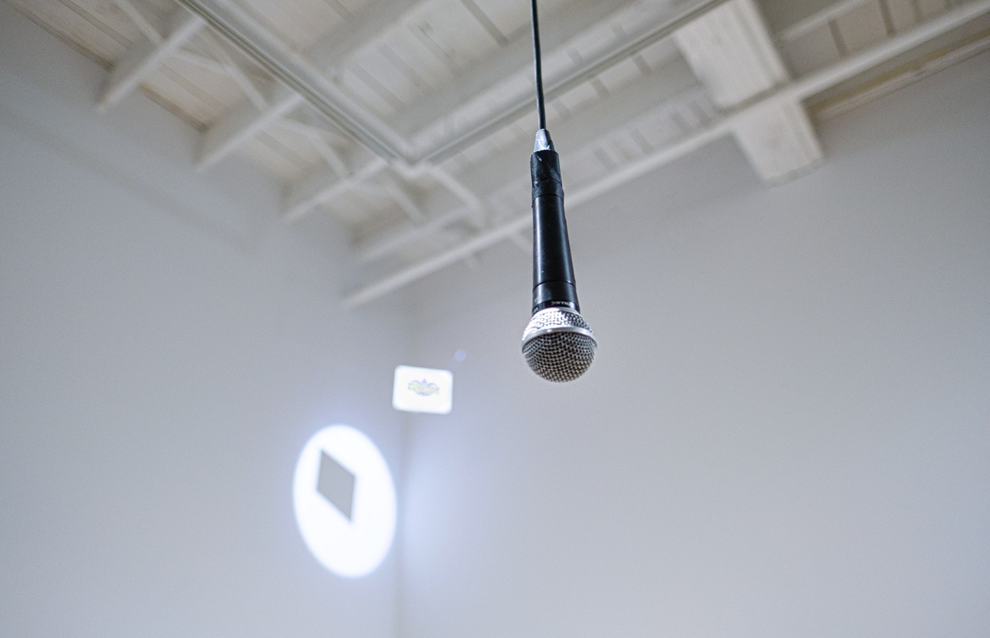 Un microphone suspendu du plafond dans une galerie blanche, avec une projection au mur dans un coin.