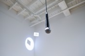Un&#x20;microphone&#x20;suspendu&#x20;du&#x20;plafond&#x20;dans&#x20;une&#x20;galerie&#x20;blanche,&#x20;avec&#x20;une&#x20;projection&#x20;au&#x20;mur&#x20;dans&#x20;un&#x20;coin.