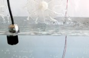 Une vue rapprochée d'un dispositif en plastique blanc ressemblant à une roue dentée ou un rotor, partiellement submergé dans l'eau. Des fils électriques noirs et rouges sont reliés à l'appareil, et des gouttelettes d'eau éclaboussent autour, suggérant un mouvement ou une action mécanique en cours.