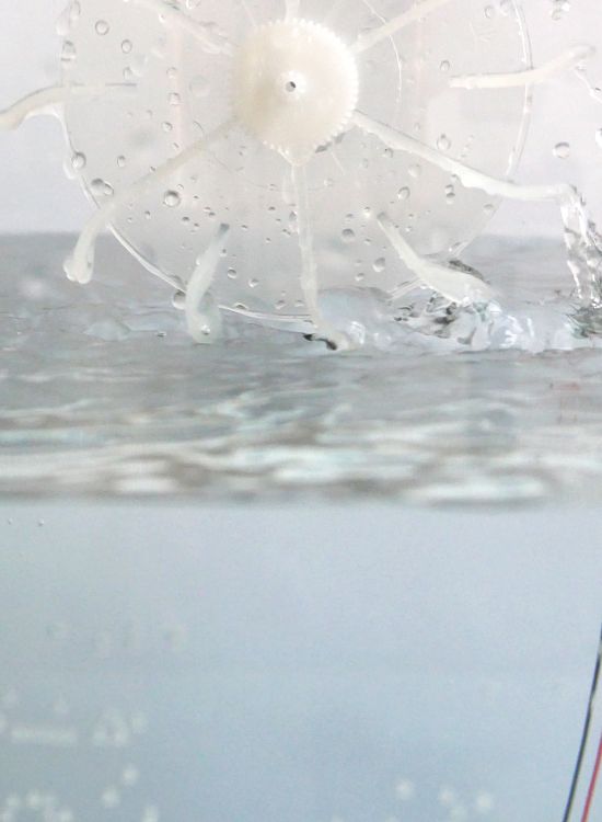 Une vue rapprochée d'un dispositif en plastique blanc ressemblant à une roue dentée ou un rotor, partiellement submergé dans l'eau. Des fils électriques noirs et rouges sont reliés à l'appareil, et des gouttelettes d'eau éclaboussent autour, suggérant un mouvement ou une action mécanique en cours.