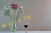 L'image montre un vase en verre contenant des fleurs diverses, principalement blanches et roses, posé sur une table blanche. À côté du vase, il y a un verre de vin rouge. On peut lire l'inscription en jaune "Les 5 à 7 de l'été" sur l’image. L'ensemble de la scène donne une impression de simplicité et d'élégance, évoquant une ambiance estivale et détendue.