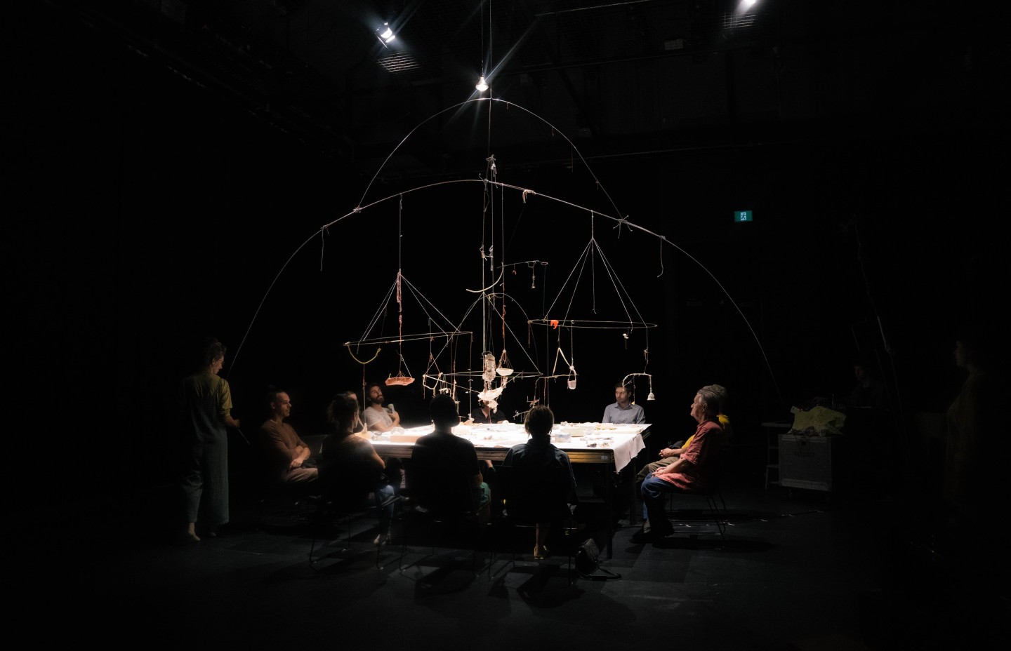 Dans une salle très sombre, une grande table rectangulaire vivement éclairée rassemble une dizaine de personnes assises autour. Au-dessus de la table, un vaste mobile composé d’arcs, de tiges et de fils suspend de petits objets, comme une constellation matérielle au centre de la scène.