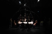 In a very dark room, a brightly lit rectangular table gathers about a dozen seated people around it. Above the table, a large suspended mobile—arcs, rods, and threads—holds small objects, forming a material constellation at the center of the scene.