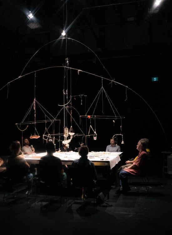 Dans une salle très sombre, une grande table rectangulaire vivement éclairée rassemble une dizaine de personnes assises autour. Au-dessus de la table, un vaste mobile composé d’arcs, de tiges et de fils suspend de petits objets, comme une constellation matérielle au centre de la scène.