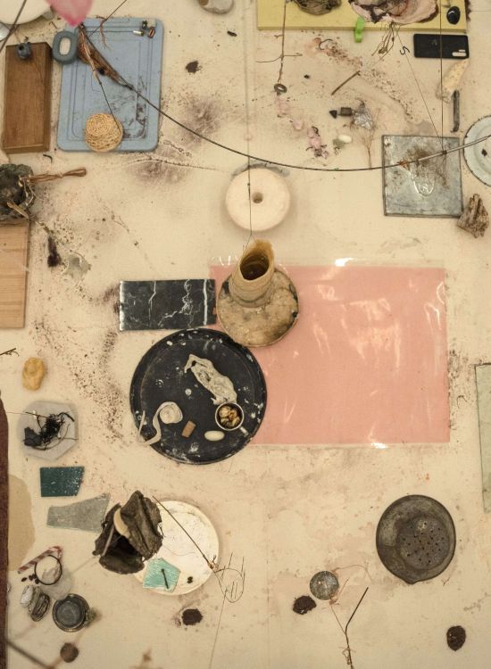 Overhead view of an installation in progress composed of fragments of materials arranged on a light surface: glass, ceramic, wood, threads, organic elements and found objects. The elements are distributed asymmetrically and connected by fine lines, suggesting a work in process and a fragile balance between materials.