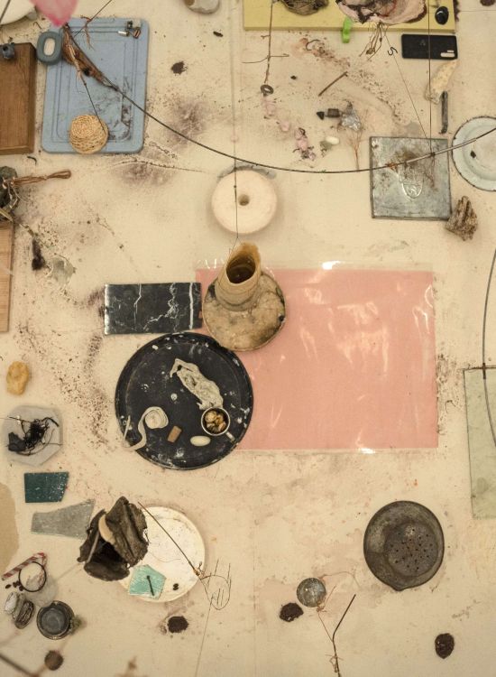 Vue de dessus d’une installation en cours composée de fragments de matériaux disposés sur une surface claire : verre, céramique, bois, fils, éléments organiques et objets trouvés. Les éléments sont répartis de manière non symétrique, reliés par des lignes fines, évoquant un processus en construction et un équilibre fragile entre les matières.