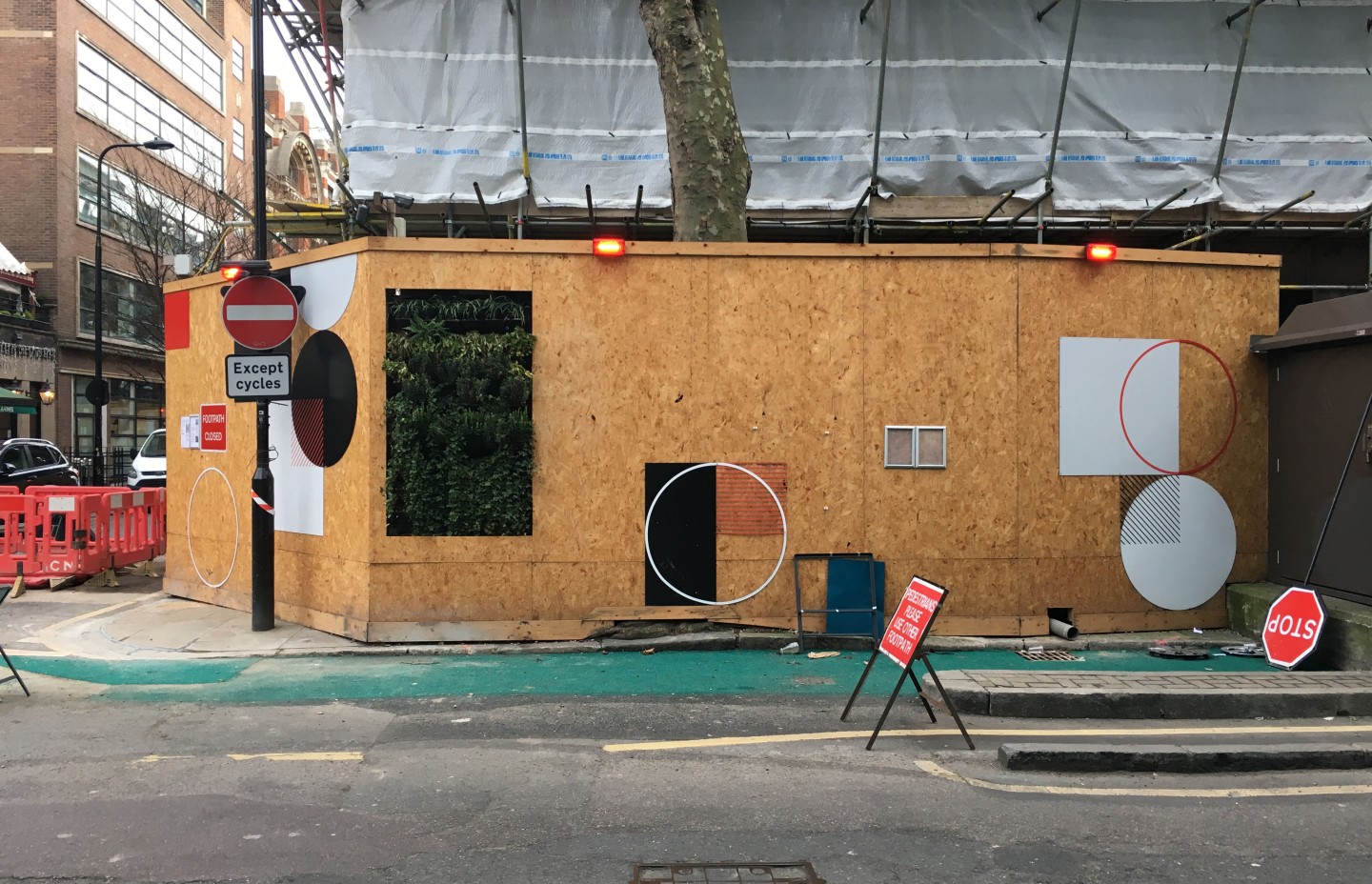 Un chantier de construction entouré de panneaux en bois avec des motifs géométriques. Des panneaux de signalisation indiquent "Except cycles" et "Fourteen closed". Il y a aussi un panneau stop renversé et un autre panneau avertissant de la fermeture de la chaussée.
